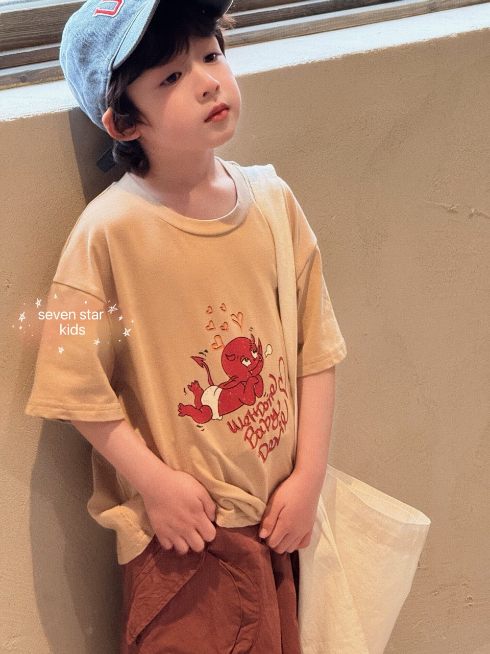 BOY wearing a beige t-shirt with a red baby devil graphic.